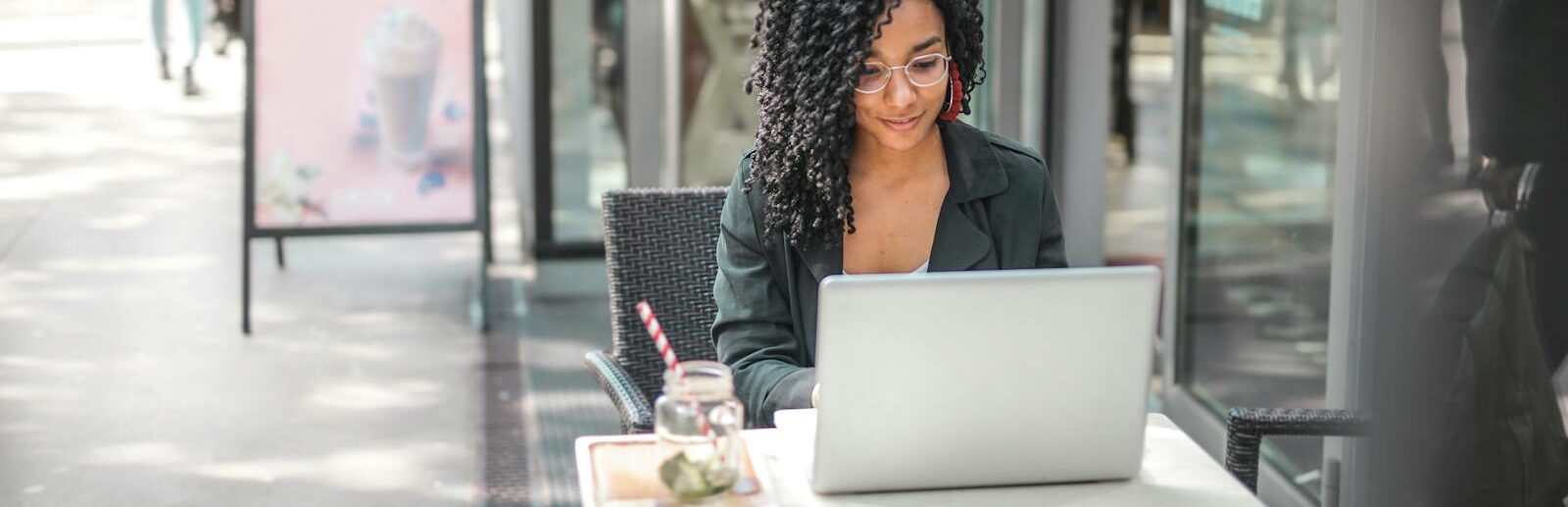 High angle of pensive African American female freelancer in glasses and casual clothes focusing on screen and interacting with netbook while sitting at table with glass of yummy drink on cafe terrace in sunny day